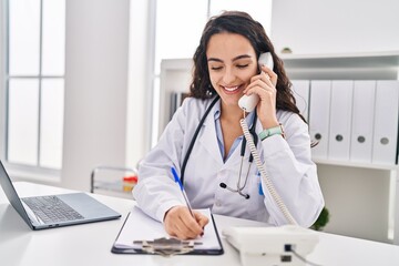 Young hispanic woman wearing doctor uniform talking on the telephone at clinic