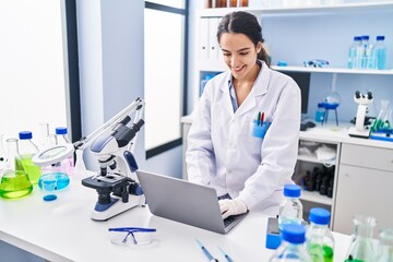 Young hispanic woman wearing scientist uniform using laptop at laboratory