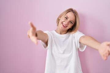 Young caucasian woman standing over pink background looking at the camera smiling with open arms for hug. cheerful expression embracing happiness.
