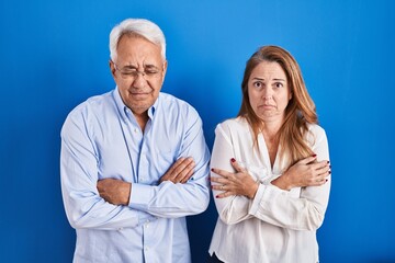 Middle age hispanic couple standing over blue background shaking and freezing for winter cold with...