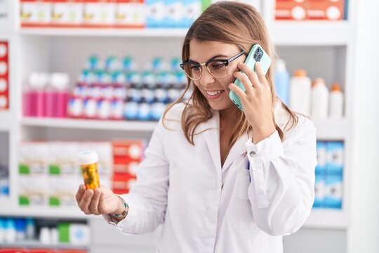 Young woman pharmacist holding pills bottle talking on smartphone at pharmacy