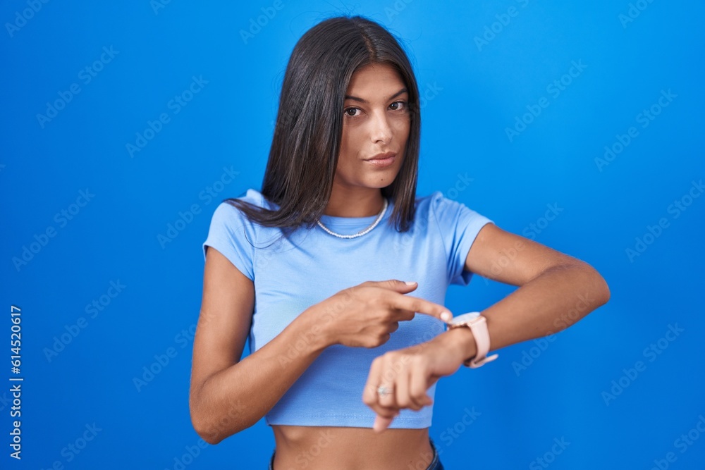 Wall mural Brunette young woman standing over blue background in hurry pointing to watch time, impatience, upset and angry for deadline delay