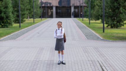 A sad caucasian girl in uniform and with a backpack holds her arms crossed on her chest stands outdoors after school.