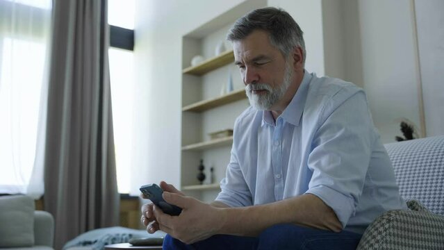 Bearded middle-aged man sitting on sofa use mobile phone smiles at home apartment. Mature man texting share messages content on smartphone social media applications online, watching relax movie