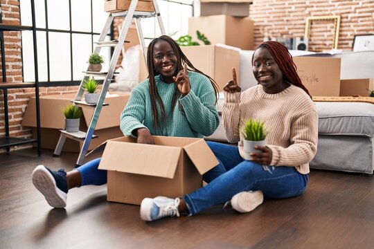 Two african woman moving to a new home sitting on the floor surprised with an idea or question pointing finger with happy face, number one - Powered by Adobe