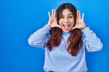 Fototapeta premium Hispanic young woman standing over blue background smiling cheerful playing peek a boo with hands showing face. surprised and exited