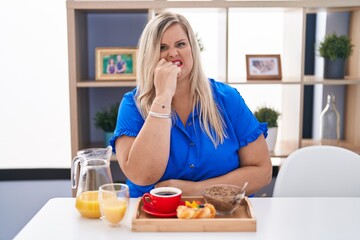 Obraz na płótnie Canvas Caucasian plus size woman eating breakfast at home looking stressed and nervous with hands on mouth biting nails. anxiety problem.