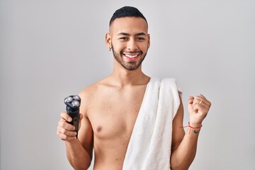 Young hispanic man holding electric razor screaming proud, celebrating victory and success very excited with raised arms - obrazy, fototapety, plakaty