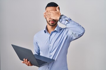 Young hispanic man working using computer laptop smiling and laughing with hand on face covering eyes for surprise. blind concept.