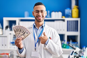 Young hispanic man working at scientist laboratory holding money pointing finger to one self...