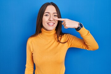 Fototapeta premium Young brunette woman standing over blue background pointing with hand finger to face and nose, smiling cheerful. beauty concept