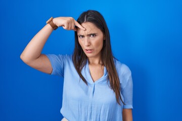 Fototapeta premium Young brunette woman standing over blue background pointing unhappy to pimple on forehead, ugly infection of blackhead. acne and skin problem