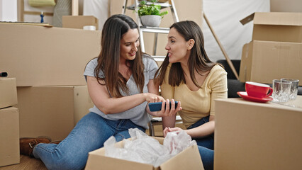 Two women using smartphone sitting on floor at new home