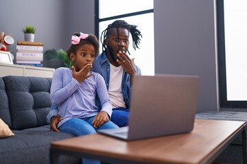 African father and daughter doing video call on laptop at home covering mouth with hand, shocked...