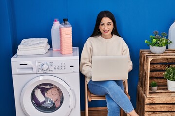 Young beautiful hispanic woman using laptop waiting for washing machine at laundry room