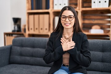 Fototapeta premium Young beautiful hispanic woman psychologist smiling confident sitting with hands on heart at psychology clinic