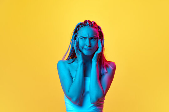 Portrait of cute, pretty young woman with stylish hairstyle posing with thoughtful grimace face against yellow studio background in neon light. Concept of youth, emotions, beauty, lifestyle, ad