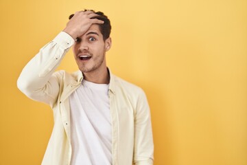 Young hispanic man standing over yellow background surprised with hand on head for mistake, remember error. forgot, bad memory concept.