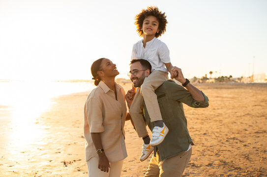 Happy young black family of three enjoying time together by seaside outdoors, father holding son on shoulders