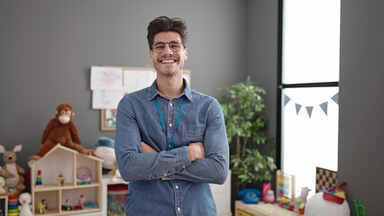 Young hispanic man preschool teacher smiling confident standing with arms crossed gesture at kindergarten