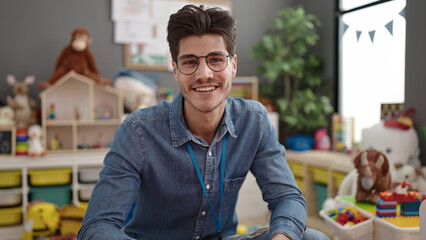 Young hispanic man preschool teacher smiling confident sitting on table at kindergarten
