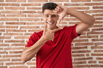 Young hispanic man standing over bricks wall smiling making frame with hands and fingers with happy face. creativity and photography concept.