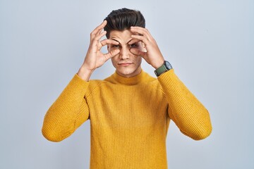 Young hispanic man standing over blue background trying to open eyes with fingers, sleepy and tired...
