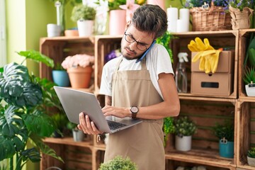 Fototapeta premium Young hispanic man florist talking on smartphone using laptop at flower shop