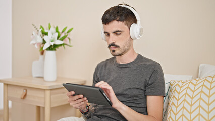 Young hispanic man using touchpad and headphones sitting on bed at bedroom