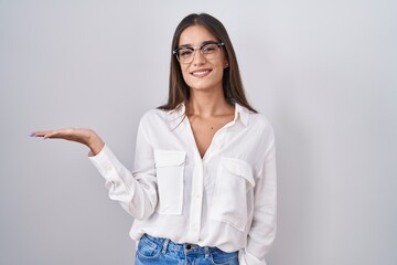 Young brunette woman wearing glasses smiling cheerful presenting and pointing with palm of hand looking at the camera.