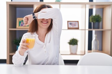 Brunette woman drinking glass of orange juice smiling cheerful playing peek a boo with hands showing face. surprised and exited