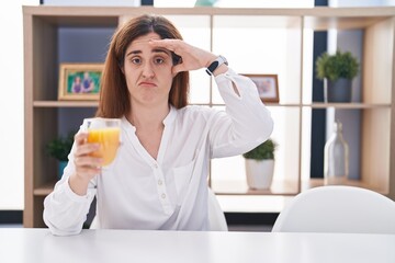 Brunette woman drinking glass of orange juice worried and stressed about a problem with hand on forehead, nervous and anxious for crisis
