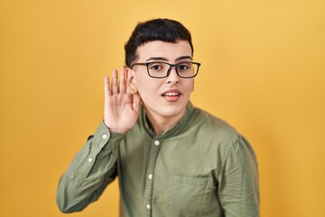 Fototapeta premium Non binary person standing over yellow background smiling with hand over ear listening an hearing to rumor or gossip. deafness concept.