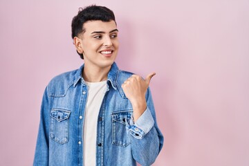 Non binary person standing over pink background smiling with happy face looking and pointing to the side with thumb up.