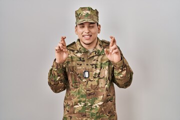 Young arab man wearing camouflage army uniform gesturing finger crossed smiling with hope and eyes...