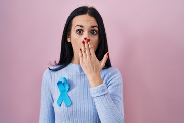 Hispanic woman wearing blue ribbon covering mouth with hand, shocked and afraid for mistake....