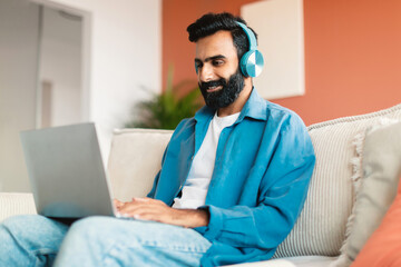 Happy indian man using computer and wireless earphones at home