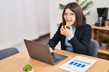 Young beautiful hispanic woman business worker using laptop eating energy cereal bar at office