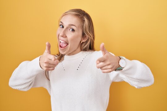 Young caucasian woman wearing white sweater over yellow background pointing fingers to camera with happy and funny face. good energy and vibes.