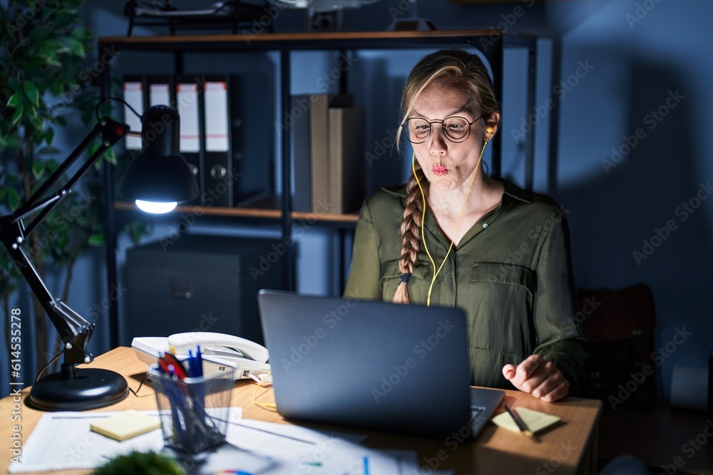 Poster Young blonde woman working at the office at night making fish face with lips, crazy and comical gesture. funny expression.