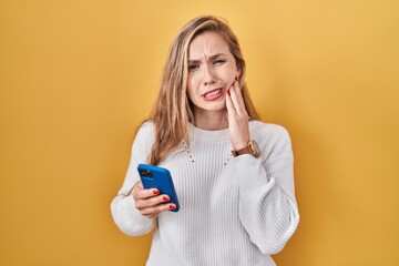 Young blonde woman using smartphone typing message touching mouth with hand with painful expression...