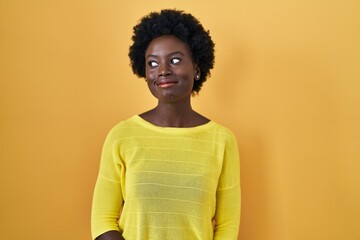 African young woman standing over yellow studio smiling looking to the side and staring away thinking.