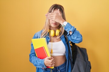 Young blonde woman wearing student backpack and holding books covering eyes with hand, looking serious and sad. sightless, hiding and rejection concept