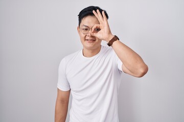 Young asian man standing over white background doing ok gesture with hand smiling, eye looking through fingers with happy face.