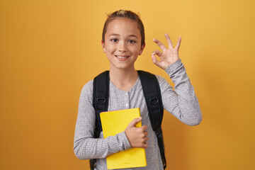 Little caucasian boy wearing student backpack and holding book doing ok sign with fingers, smiling friendly gesturing excellent symbol