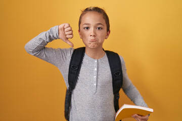 Little caucasian boy wearing student backpack and holding book with angry face, negative sign...
