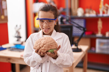 Fototapeta premium Little hispanic girl holding brain at science class at school sticking tongue out happy with funny expression.