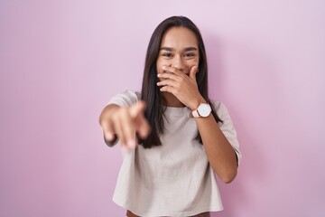 Young hispanic woman standing over pink background laughing at you, pointing finger to the camera...