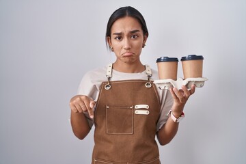 Young hispanic woman wearing professional waitress apron holding coffee pointing down looking sad...