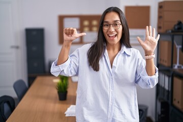 Plakat Young hispanic woman at the office showing and pointing up with fingers number six while smiling confident and happy.